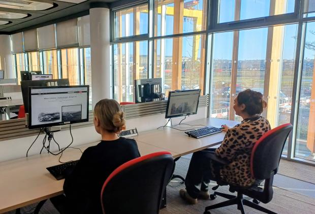 Image of two women sitting at a large desk in front of computer screens in an office room. The room has floor to ceiling windows and a yellow crane against a blue sky is visible.
