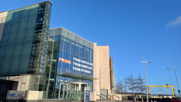 Image of a large stone and glass building on a sunny day. A banner on the front of the building reads 'PRONI: Your Archives, Your Heritage, Your Stories' and a yellow crane is visible in the right hand side of the image.