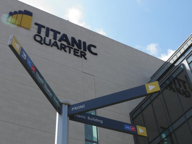 Image of a wall of a large grey building with the words 'Titanic Quarter' with three street signs on a post in the foreground.