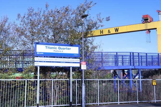 Image of a train station platform and footbridge on a sunny day. A sign reads 'Titanic Quarter, Platform 2' with additional signage underneath for directions. A large yellow crane with letters in black reading 'H&W' is visible on the right hand side.
