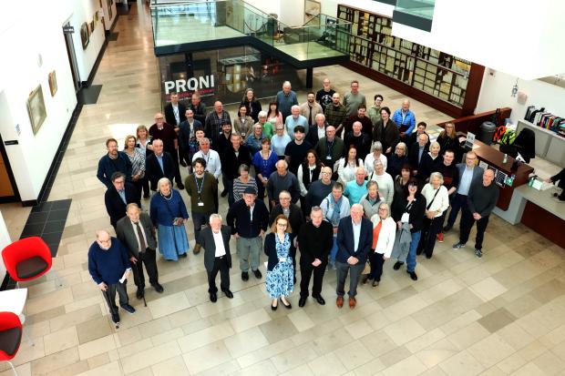 A large group of around 50 people stand in the PRONI atrium