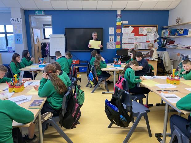 a classroom of school children sitting at their desks while a man at the top of the room holds a large yellow page