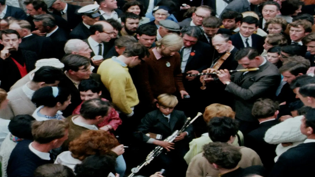 Colour photograph of a Fleadh session in the 1960s with a large crowd on a public street. A young boy sits in the middle of the crowd playing the uillean pipes with other men around him playing the flute and the fiddle.