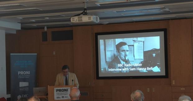 A man in a cream suit stands at the podium in the PRONI lecture theatre. Behind him on the screen is a black and white photograph of Sam Hanna Bell smoking a pipe at his desk. The text on the screen reads 'BBC Radio Ulster - Interview with Sam Hanna Bell'
