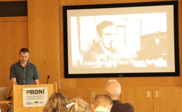 A man in a blue shirt speaking at the podium in the PRONI lecture theatre. Behind him on the screen is a black and white photograph of Sam Hanna Bell smoking a pipe at his desk. The text on the image reads 'BBC Radio Ulster - Interview with Sam Hanna Bell'