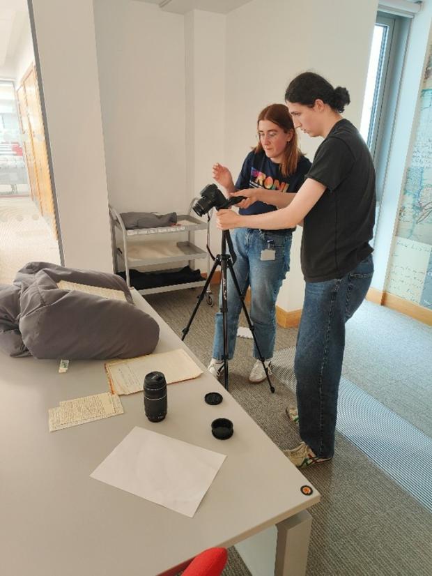 Two individuals stand behind a camera on a tripod, the camera is pointed towards some original documents on a book cushion.