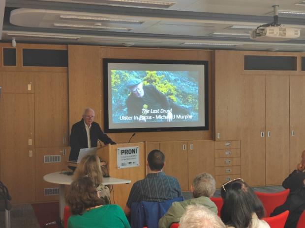 A man in a white shirt and black jacket standing behind the podium in the PRONI lecture theatre. Behind him on the screen is a colour still from the film showing Michael J Murphy lying in from of a gorse bush. The text on the image reads 'The Last Druid - Ulster In Focus - Michael J Murphy'