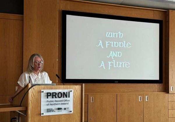 A blonde woman with glasses stands at the podium in the PRONI lecture theatre. Behind her on the screen is a title card that reads 'With a fiddle and a flute'