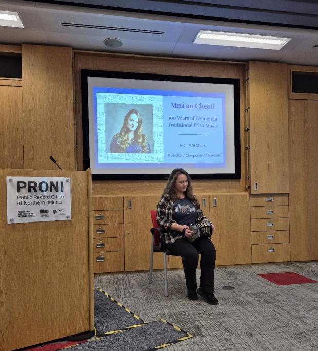 A woman with long brown hair sitting on a chair in the PRONI lecture theatre playing the concertina