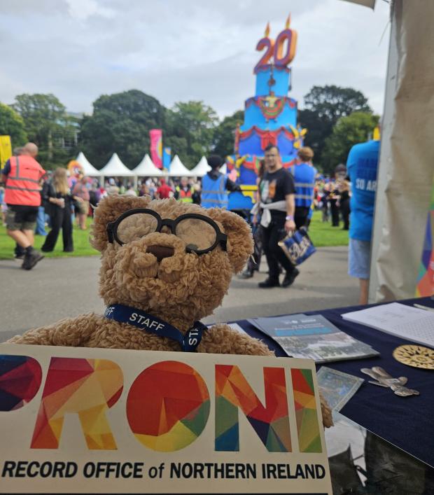 A toy teddy bear wearing glasses and a staff lanyard holds a sign that reads 'PRONI Public Record Office of Northern Ireland'. In the background, there is a large float from the parade, decorated as a blue cake with candles that read '20' on top.