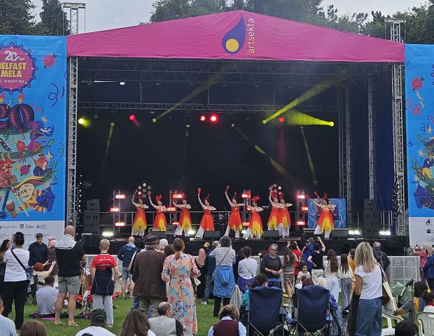 Image of a stage set up in a park. On the stage are a group of dancers dressed in orange and yellow dresses. All of the dancers are holding a red fan in the air with their right arms