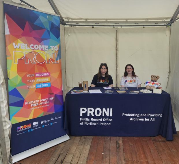Two women with dark hair sitting behind a table covered with a navy tablecloth that reads 'PRONI Public Record Office of Northern Ireland' - To the left-hand side of the table is a colourful pop-up banner that reads 'Welcome to PRONI'