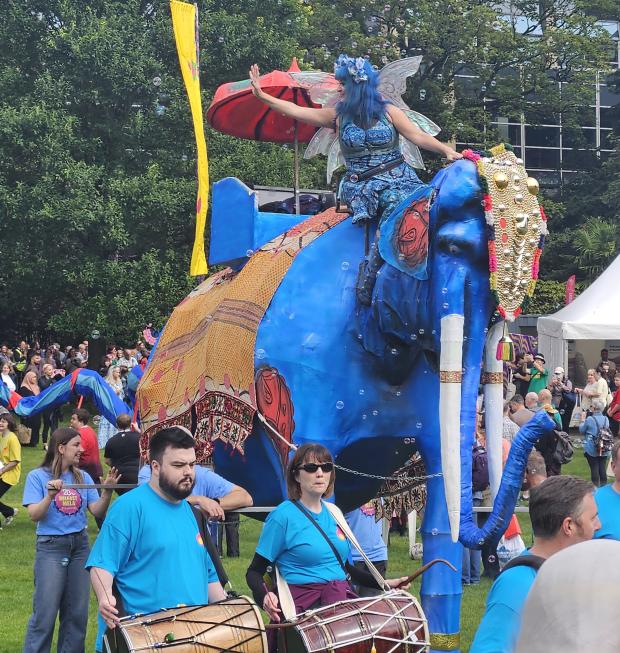 A woman dressed as a fairy with a blue outfit and hair sits on top of a float decorated as a large blue elephant. She is waving to the crowd and there are bubbles floating around her. In front of the elephant are two individuals playing large barrel drums