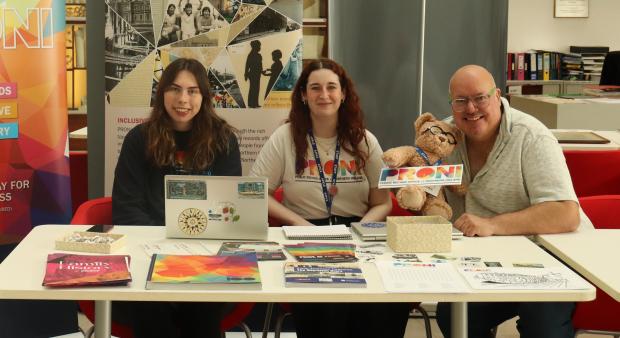 Three people sitting behind a table, one is holding a teddy bear with glasses. On the table are a laptop and promotional leaflets for PRONI.