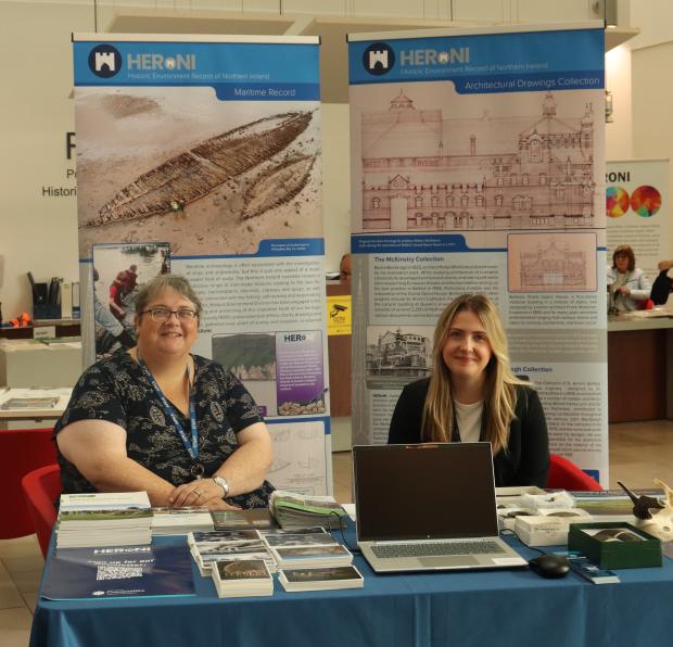 Two people sitting behind a table with a blue tablecloth on it - Behind them are two pop-up banners advertising HERoNI