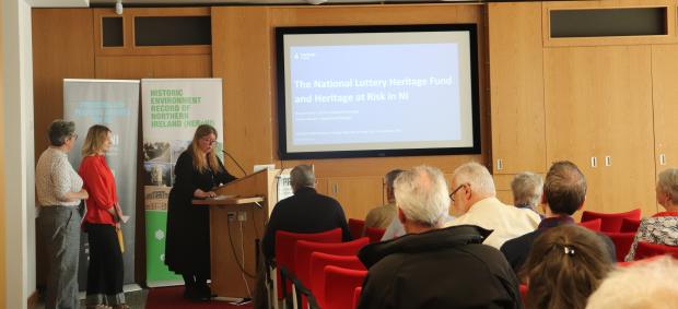 three women stand behind a podium at the top of a conference room in front of audience members. Behind them on the screen is a presentation with a blue background on the subject of the National Lottery Heritage Fund