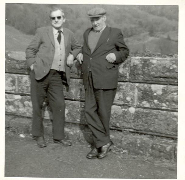 Black and white image of two men leaning on a wall with trees in the background. The man on the left is wearing sunglasses and the man on the right is wearing a cap.